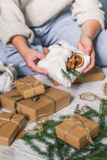 A person holding a gift wrapped in fabric decorated with a dried orange slice, cinnamon sticks and sprigs of pine
