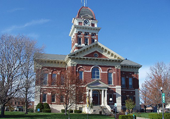 Two-story red brick building featuring a four-stage, square clock tower with a pyramidal slate roof atop the intersecting wings