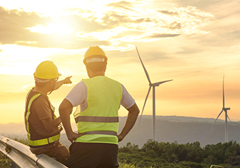 Two men wearing hard hats and safety vests look at a wind turbine. One man is pointing. The background is a morning sky with hills behind the turbine.  A forest sits between the men and the turbine.