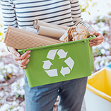 A woman holding a green tub with the recycling logo, standing next to three recycling bins for separating recyclables