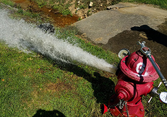 A red fire hydrant with gauges attached being flushed, releasing a large amount of water on a grassy area