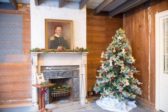 Decorated Christmas tree in the corner of room near fireplace with portrait of a gentleman dressed in a military uniform hangs above the mantle