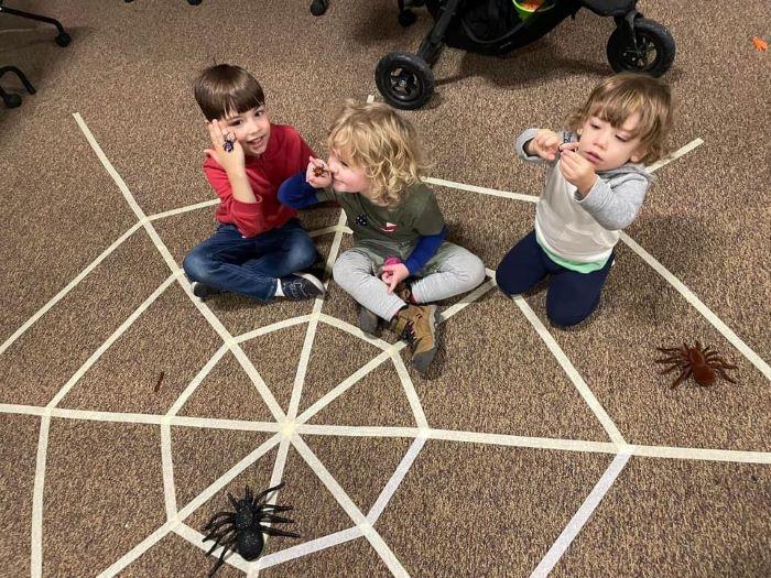 Three toddlers sitting on the floor in a spider web taped to the carpet, playing with fake spiders.