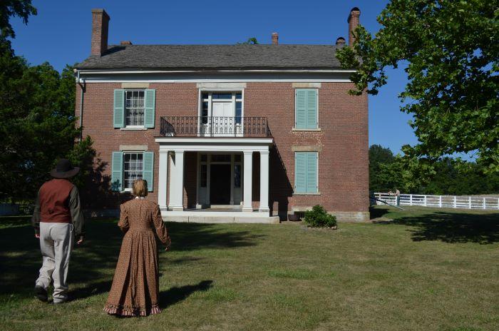 Man and woman dressed in period clothing standing in front of a two-story brick house.