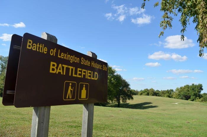Brown sign with words “Battle of Lexington State Historic Site Battlefield” and graphics depicting a person hiking. In background are trees on hill.