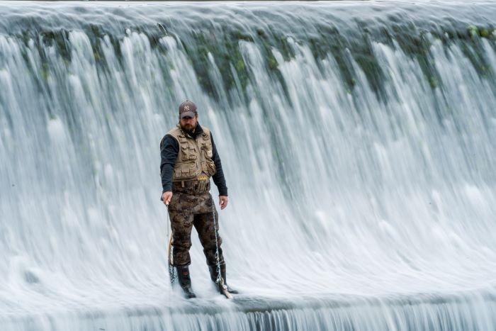 Angler standing in flowing water wearing camo waders, a fly fishing vest and blue shirt with gray hat, holding a fly fishing rod in his right hand