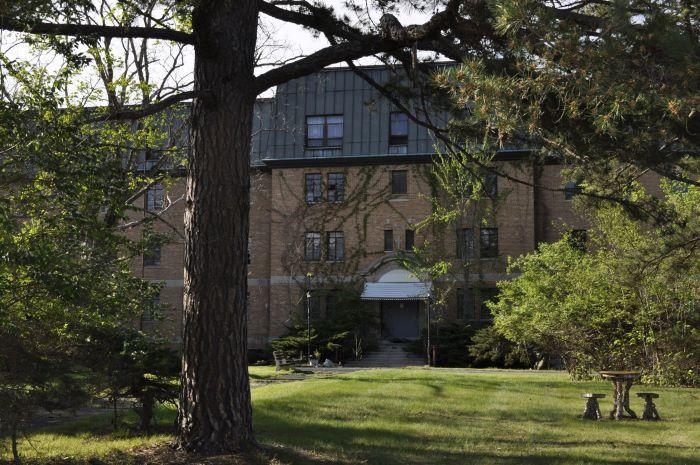 Multi-story light colored brick building with white covered entrance, metal roof and surrounded by trees.