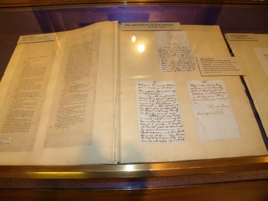 The original The Adventures of Tom Sawyer manuscript opened to display hand written text. Manuscripts is displayed in a glass case.