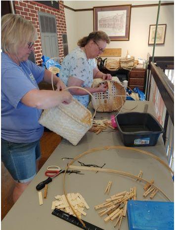 Woman in blue shirt is weaving a basket; woman in a white shirt with a pattern standing in the background also weaving a basket.