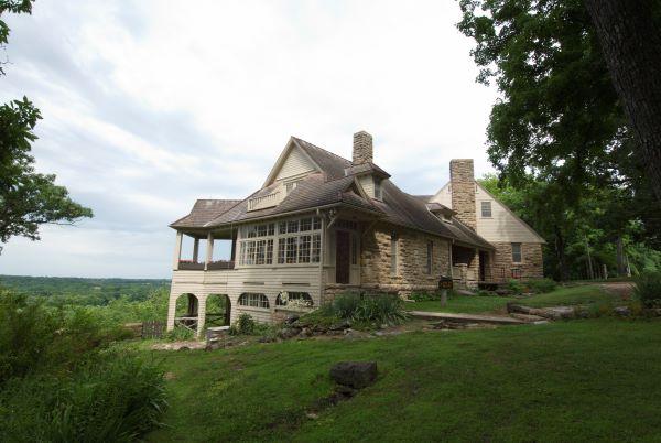 Bothwell Lodge, a historic stone house with a wraparound porch, sits on a lush green hill overlooking a scenic landscape under a partly cloudy sky.