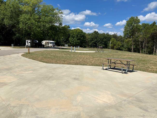 Campgrounds at Babler SP with paved sites, green grass, mature trees, picnic tables, and RVs parked at campsites under a blue sky.