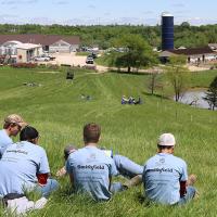 Four students sit on a hillside looking down at a field, a pond and several buildings at Carver Farm.