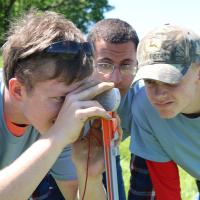 Three students use a distance measuring tool on a tripod. One boy looks through the sight while another boy takes the readings on the side of the sighting device.