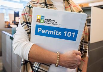 A department staff member, holding a 3-ring binder that states "Permits 101"