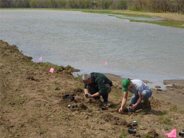 Two men dig to put in plants on the bank of water in a wetland.