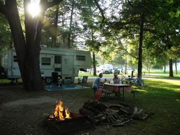 Park visitors sit at a picnic table near a campfire in front of their camper. Lawn chairs and bicycles sit nearby.