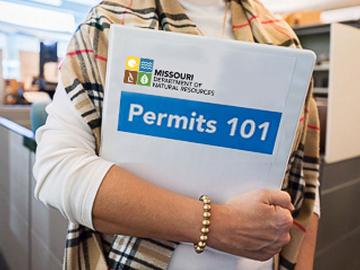 A department staff member, holding a 3-ring binder that states "Permits 101"