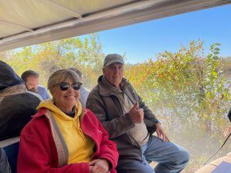 A smiling woman and a man shows his approval with a thumbs up while riding on the tram, enjoying the fall colors of the Katy Trail.