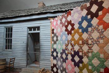 Quilt with multicolor octagonal design hangs next to a white historic building at Salt River Quilt Show.