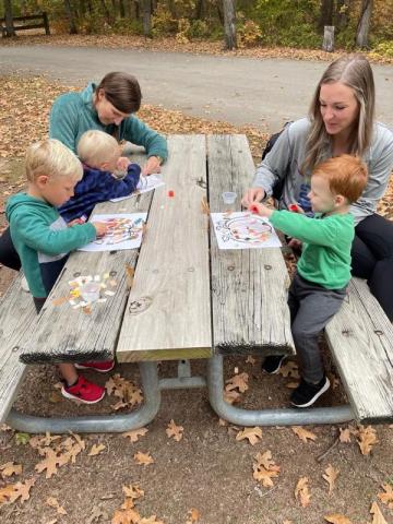 Three toddlers and two adults sit at a picnic table creating crafts. 