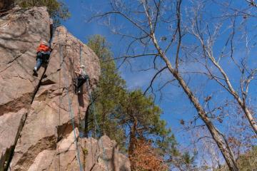 One person dressed in orange shirt, white helmet and blue jeans is climbing up the rocks assisted with blue ropes. On the right another person in a t-shirt, blue jeans and white helmet is attempting to reach the top of the rocks using blue ropes as assistance.