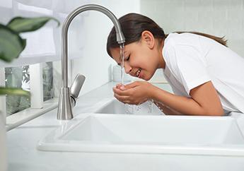 A young girl with her hands together over a sink bringing water from a running kitchen faucet to her mouth for a drink