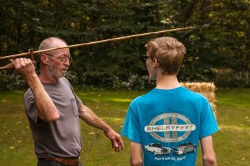 Gentleman in gray shirt holding atlatl, demonstrating how to use to younger male in blue t-shirt.