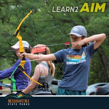 3 young girls aiming compound bows ready to fire. One in blue cap and shirt, one in orange cap with gray top and one in white cap and blue shirt.
