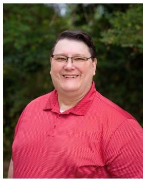 A smiling Eric Medlock with short dark hair and glasses, wearing a red polo shirt, outdoors with green trees in the background.