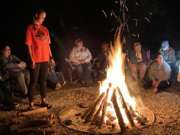 People gathered around a large, crackling campfire at night, with one person standing near the flames and others seated in a circle.