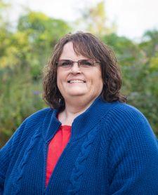Rosie Schulte, with glasses smiling outdoors, wearing a blue sweater and red shirt, with green foliage in the background.