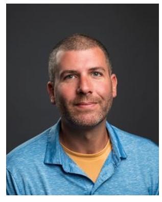 Fletcher Bone, with short hair wearing a blue collared shirt over a yellow t-shirt, posed against a plain dark background.