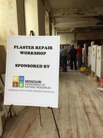 A sign advertising a "Plaster Repair Workshop" sponsored by the Missouri Department of Natural Resources and the Missouri State Historic Preservation Office, with people and workshop materials visible in the background.