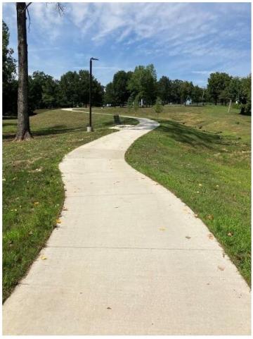 Curving concrete walkway through grassy park with trees, a bench, and a lamp post under a partly cloudy blue sky