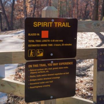 A brown sign with Spirit Trail at the top in front of a snowy ground and forest.