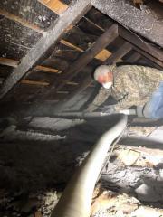 A man in an attic is wearing a face mask sprays insulation through a hose.