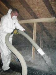 A man in an attic is wearing a hazmat suit and mask sprays insulation through a hose.
